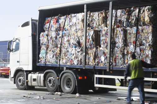 A large side-view of a white commercial cargo truck loaded with tightly packed compressed recyclable paper and cardboard waste, secured with black straps. The materials display a mixture of white, brown, and coloured papers with visible textures ranging from smooth to crinkled. The truck is parked outdoors on a paved surface, possibly in a recycling or waste disposal area near Newham. The surrounding environment includes a blurred background with some vehicles and a person in a high-visibility yellow vest walking alongside the truck, indicating active waste collection or removal. The scene emphasizes the nature of rubbish removal services in London’s East End, with the focus on the vehicle’s load of paper waste awaiting transport to recycling facilities, illustrating the work of Office Clearance Newham in managing refuse collection and disposal.