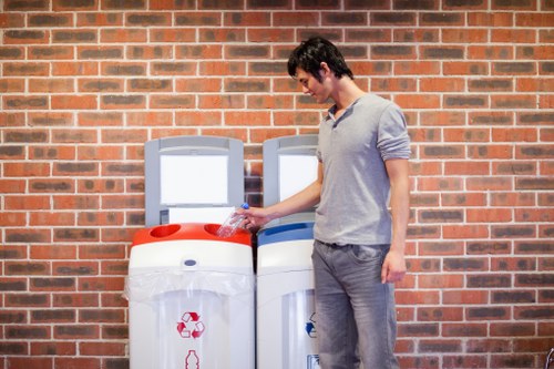 A young man with dark hair, dressed in a light grey long-sleeved shirt and grey pants, standing on a paved surface beside two white recycling bins with red and blue lids, positioned against a brick wall. He is holding a crumpled plastic bottle, appearing to place it into the recycling bin. The bins are situated outdoors, likely on a residential or commercial property in Newham, with the brick wall providing a textured background. The scene depicts a typical waste disposal activity related to rubbish management services offered by Office Clearance Newham, emphasizing the proper separation of recyclable waste materials. The environment is brightly lit, with the focus on the individual's action of discarding plastic waste responsibly into designated bins, aligning with local waste collection and recycling efforts in the area.