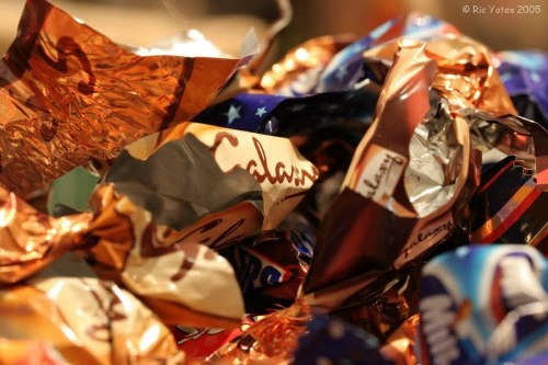 A close-up view of a pile of crumpled, metallic foil wrappers and plastic packaging, featuring various colors including copper, silver, blue, and white, with some prints and logos partially visible. The wrappers are piled together in a chaotic manner, with some edges curled and overlapping, creating a textured surface. The background appears blurred, suggesting the wrappers are on a surface such as a pavement, driveway, or indoor flooring, consistent with waste or rubbish collected for removal. The lighting is warm and diffuse, highlighting the reflective surfaces of the wrappers and emphasizing their textures. This image is representative of waste materials that might be collected by a rubbish removal service, such as those provided by Office Clearance Newham, serving areas including Newham in London.