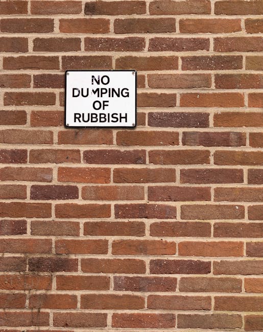 A rectangular white metal sign with bold black lettering mounted on a red brick wall. The sign reads 'NO DUMPING OF RUBBISH,' with the words arranged in three lines. The brick wall has a traditional r