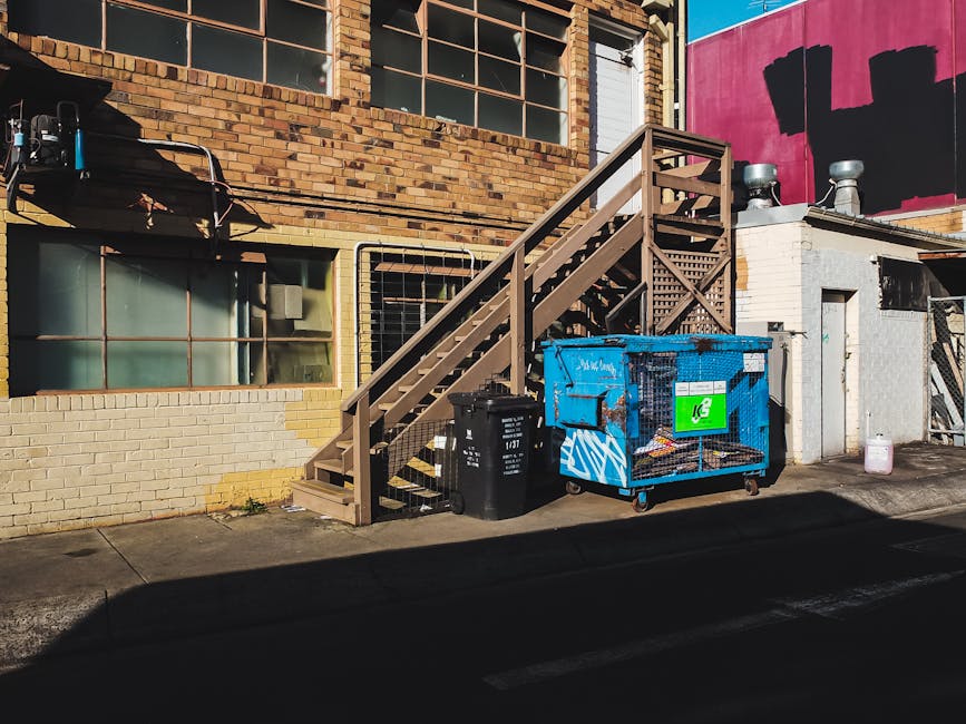 Two green wheeled rubbish bins are positioned side by side on a paved surface with a low brick border behind them. The bin on the left has a flat, slightly textured surface with a white circular label