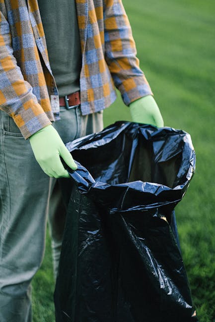 A person wearing a yellow and blue checked shirt over a dark green top, with beige trousers and a brown belt, is standing outdoors on a grassy area. They are holding open a large black rubbish bag lin