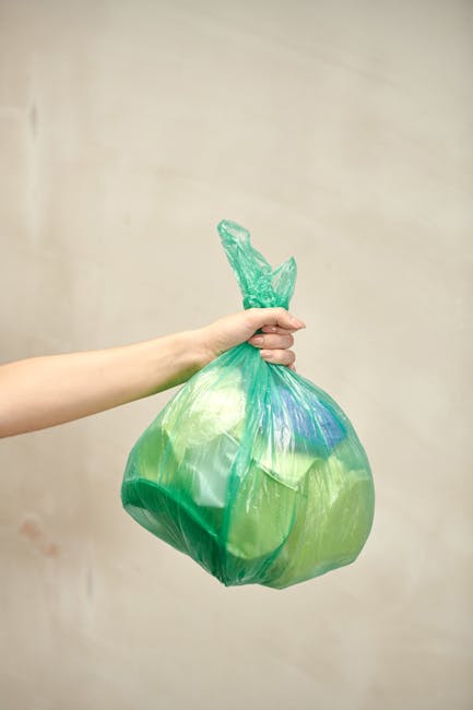 A person wearing a yellow and blue checked shirt over a dark green top, with beige trousers and a brown belt, is standing outdoors on a grassy area. They are holding open a large black rubbish bag lin